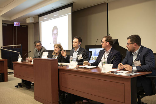Five seated panelists and a virtual participant seen on a screen in the background discuss the evolution of instant payments during the 2024 Chicago Payments Symposium.